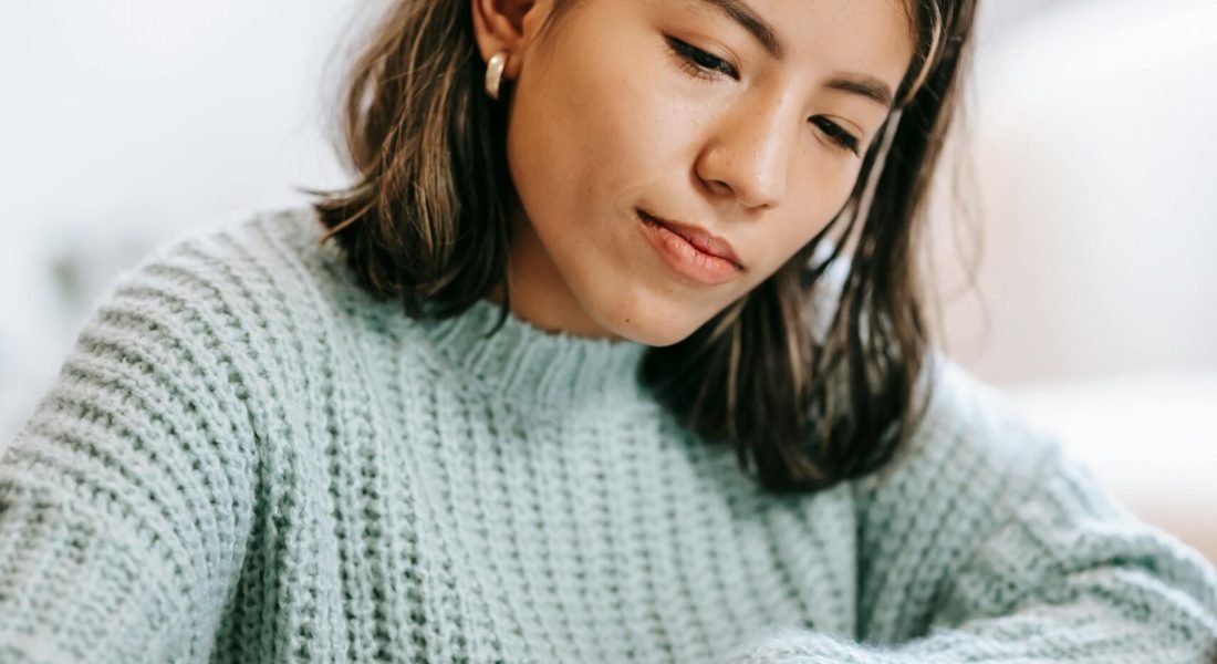 Young Hispanic female student in casual outfit using laptop while taking notes in notepad with pen at table in light apartment